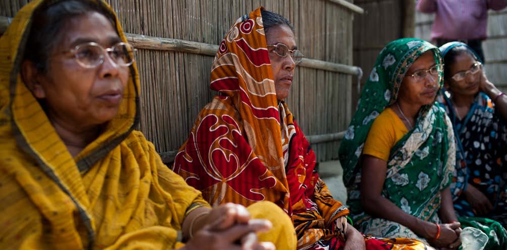 Four women in traditional South Asian attire sit together outdoors, attentively observing something off-camera.