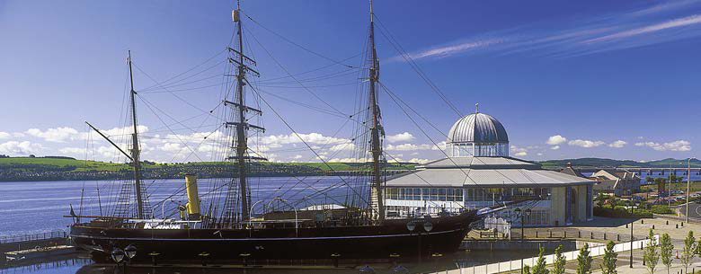 A large historical sailing ship docked next to a modern building with a domed roof and a waterfront promenade.