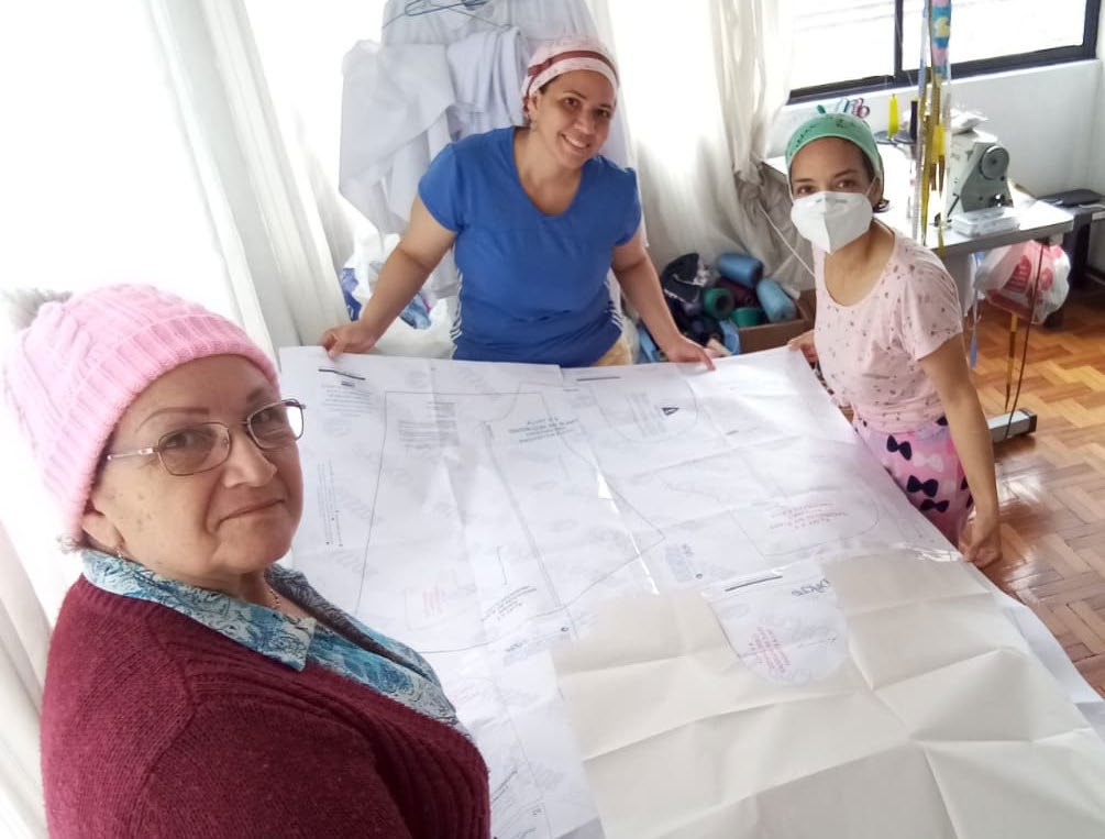 Three women looking at a large paper pattern on the floor, likely for cutting fabric, in a bright room.