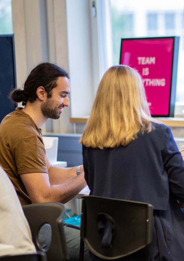 A man smiling while working at a desk next to a woman whose back is to the camera, with a pink sign in the background reading "TEAM IS EVERYTHING".