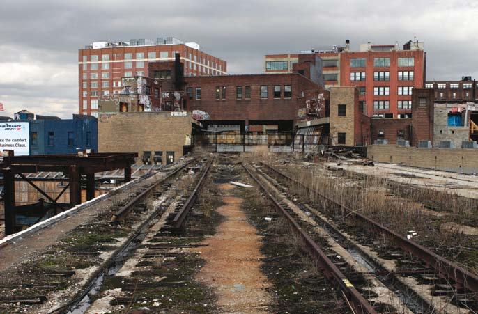 Overgrown, disused railroad tracks in the foreground leading towards a mix of residential and industrial buildings under a cloudy sky.