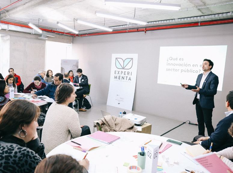 A presenter standing in front of a screen displaying "¿Qué es innovación en el sector público?" to an audience seated at round tables.