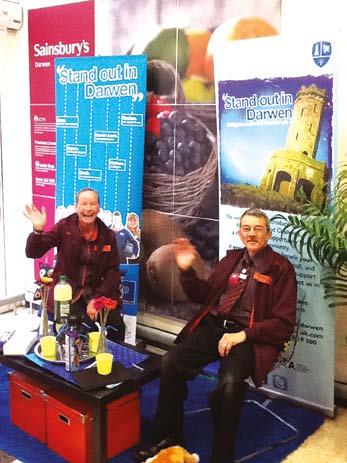 Two men in red shirts wave beside "Sainsbury's Darwen" banners promoting the "Stand out in Darwen" initiative, one with a graphic.