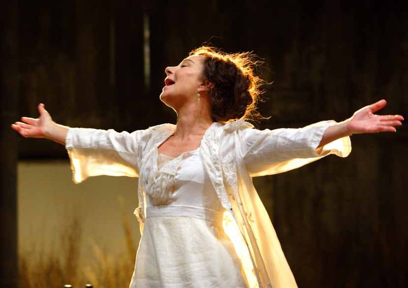 Woman in a white dress with outstretched arms and head tilted back, singing or expressing strong emotion on stage.