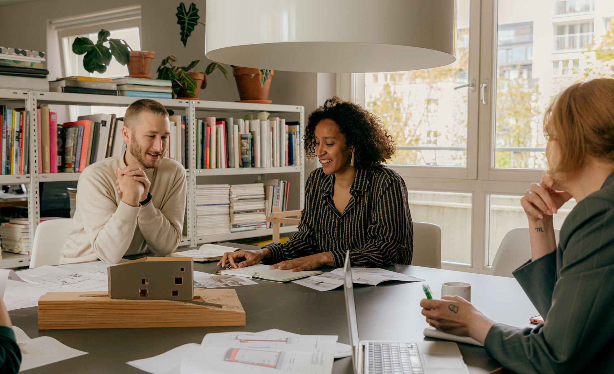 Three architects discussing a building model on a table with blueprints and laptops.