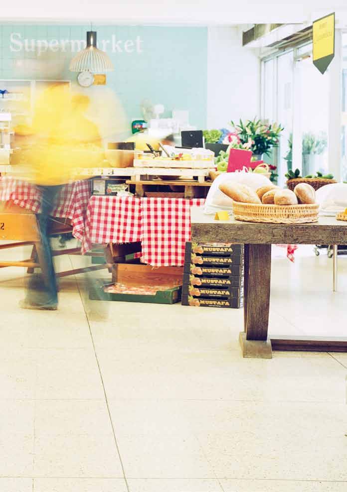 Interior of a supermarket with a blurred person, a table displaying fresh bread, and other produce stands.