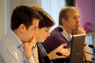 Three professionals sitting at a table with microphones and a laptop, appearing engaged in a meeting or conference.
