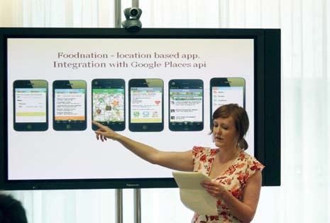 Woman presenting a "Foodnation" location-based app, pointing at a large screen displaying app screenshots and text about Google Places API integration.