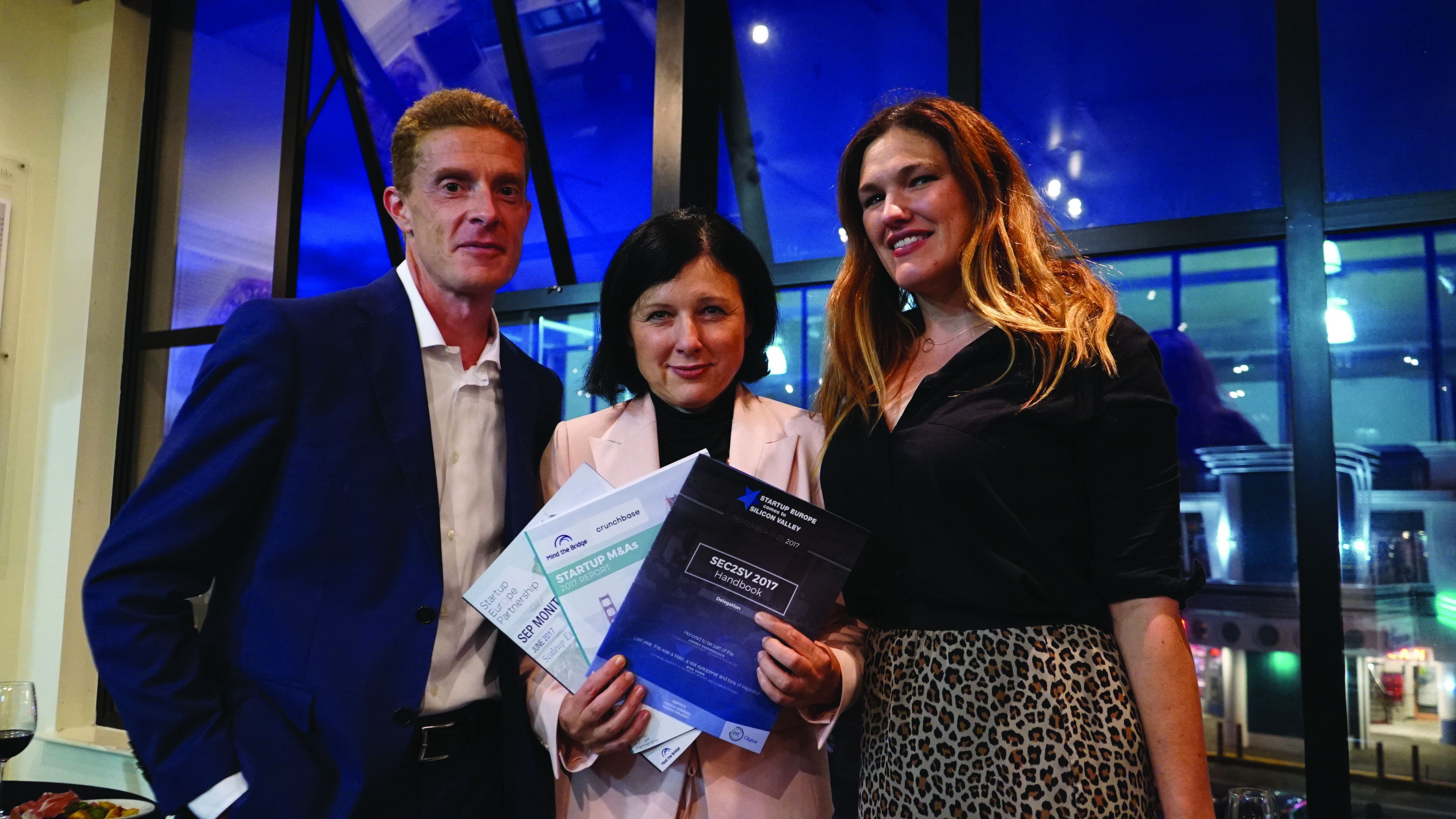 Three professionals, two women and one man, stand smiling while holding reports titled "STARTUP MAAS" and "SECSEV 2017" at an event.