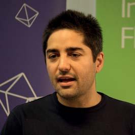 Close-up portrait of a man with dark hair and a beard, speaking, with a "Nesta: Making Innovation Flourish" banner in the background.