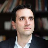 Headshot of a man with dark hair and a beard, looking to the right, with bookshelves in the background.