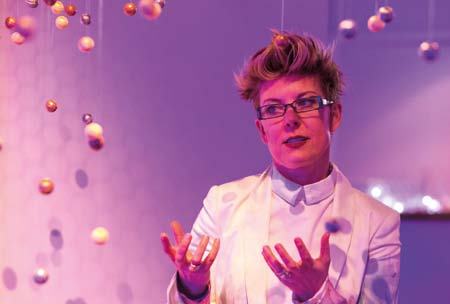 A woman with spiky hair and glasses, dressed in a white blazer, gestures with open hands surrounded by suspended, glowing spherical objects, suggesting dynamic interaction or conceptual complexity.