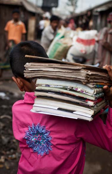 A young person, viewed from behind, carries a stack of books and papers on their shoulder through a bustling outdoor area.