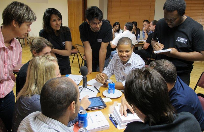 A diverse group of people engaged in a collaborative meeting or workshop around a table with a microphone, tablet, and papers.