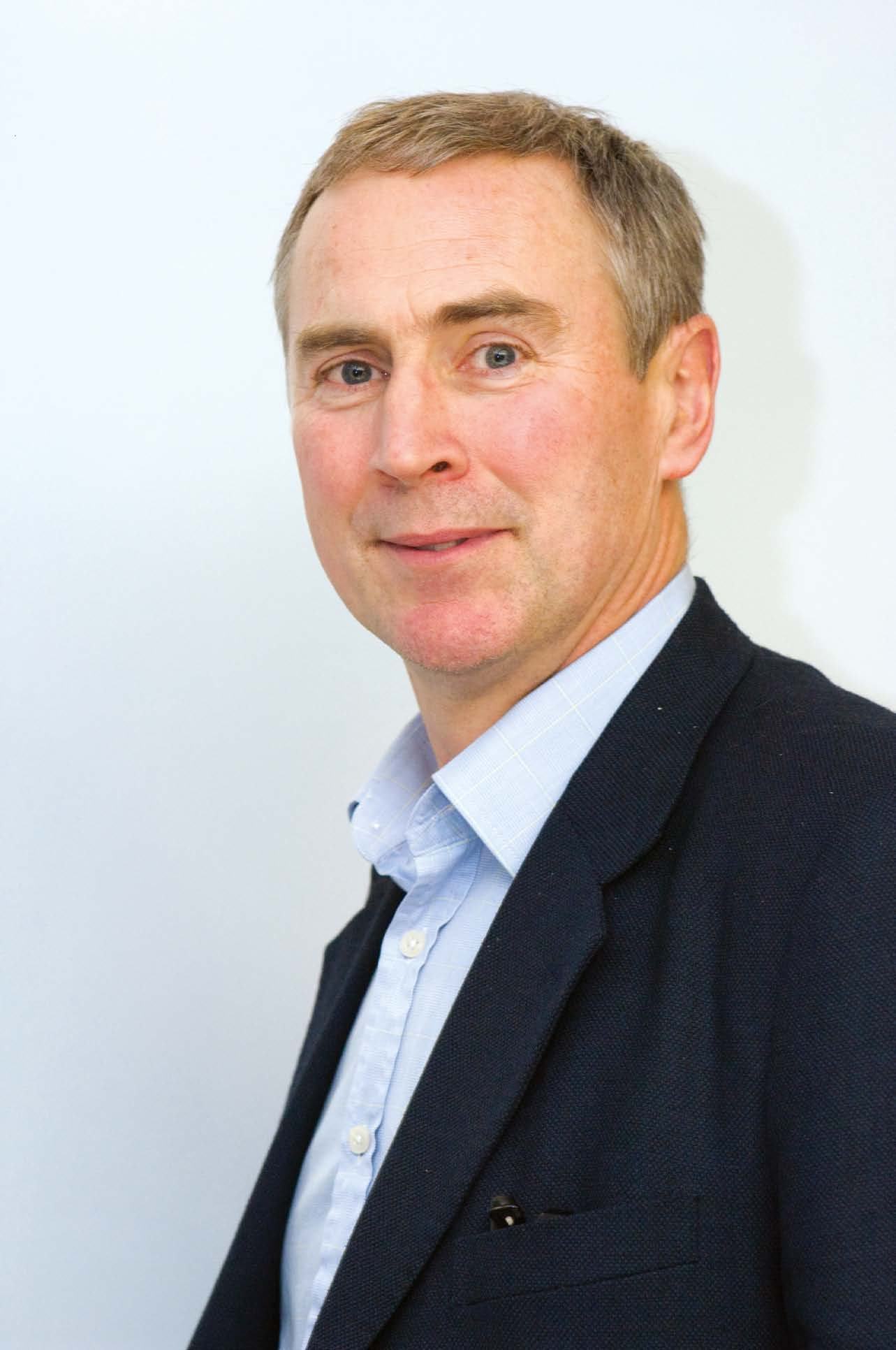 Portrait of a man with grey hair, blue eyes, wearing a blue shirt and dark blazer.