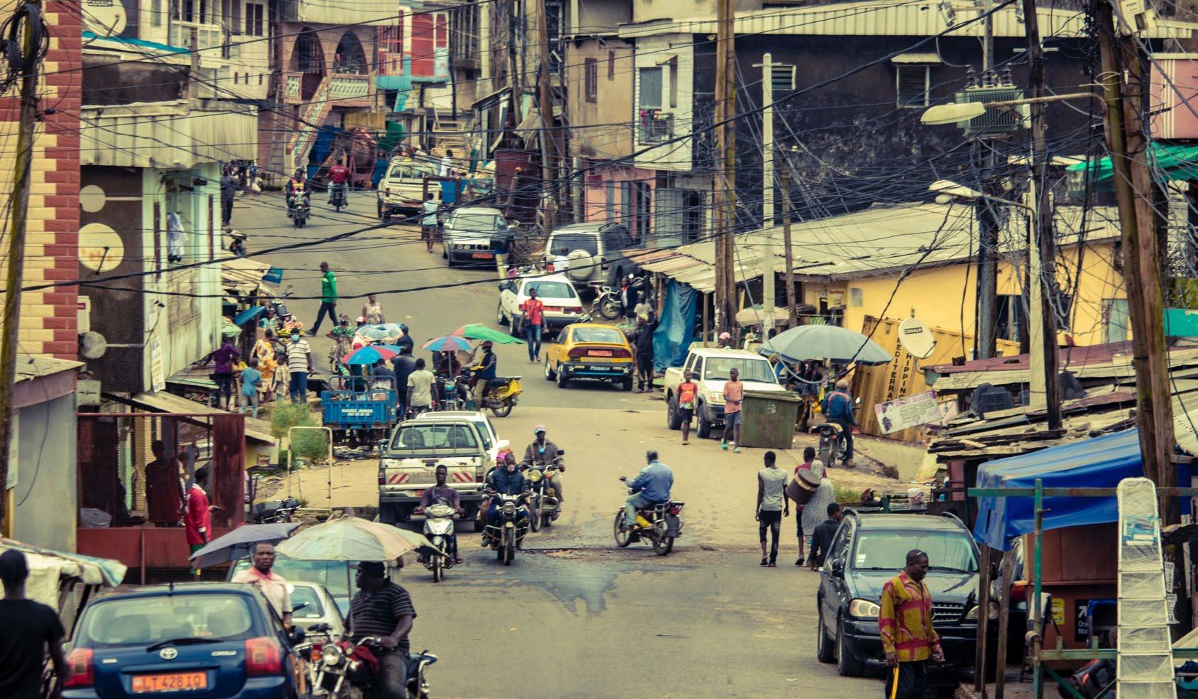 A bustling street scene in a city, with numerous pedestrians, vehicles, buildings, and overhead power lines, depicting urban life.