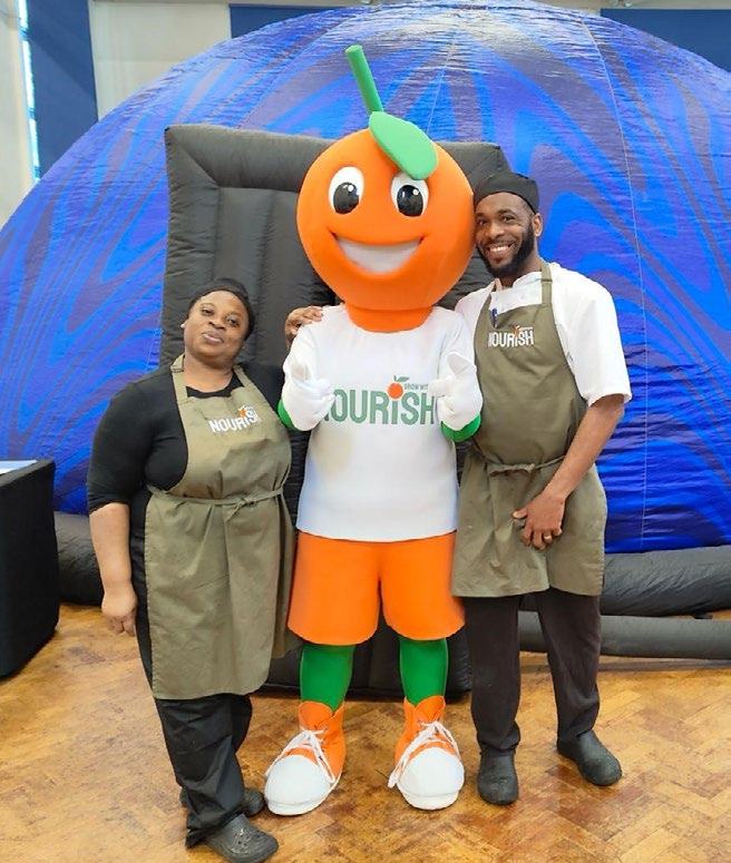 Two staff members in 'Nourish' aprons stand with an orange fruit mascot wearing a 'Grow It Nourish It' t-shirt in front of a blue inflatable dome, likely at an event.