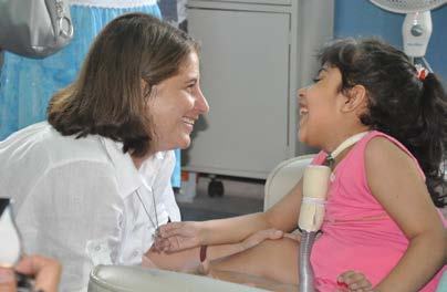 A woman in a white shirt smiles at a laughing young girl in a pink top in what appears to be a care setting.
