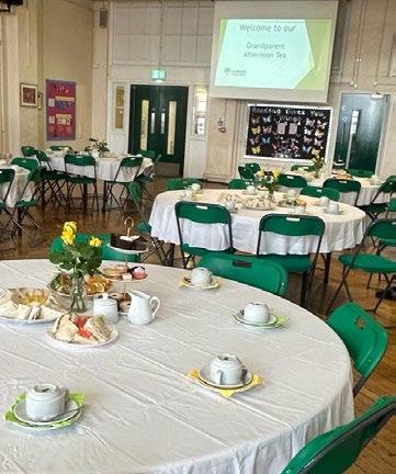 A room filled with round tables set for a "Grandparent Afternoon Tea," with teacups, sandwiches, and cakes, and a projector screen displaying the event title.