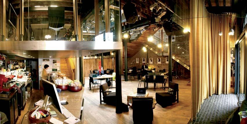 Wide shot of a modern, multi-level interior space with exposed brick, wooden floors, tables, chairs, and people working.