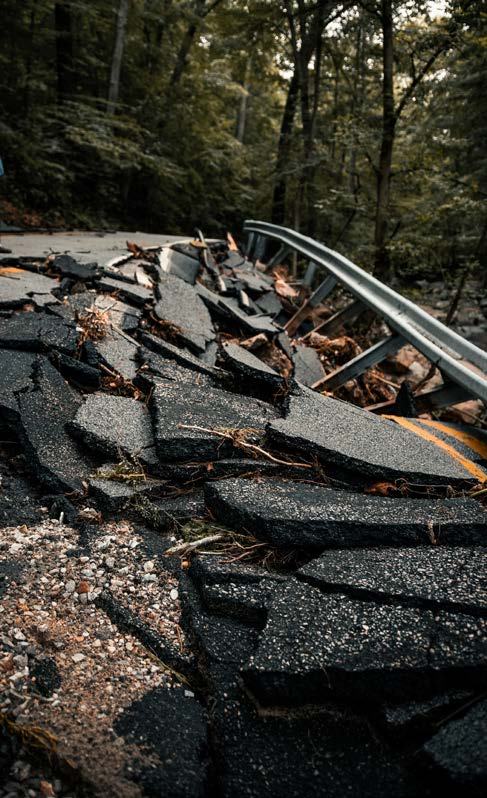Severely damaged asphalt road, broken into large chunks, with a bent guardrail, indicating infrastructure failure or natural disaster impact.