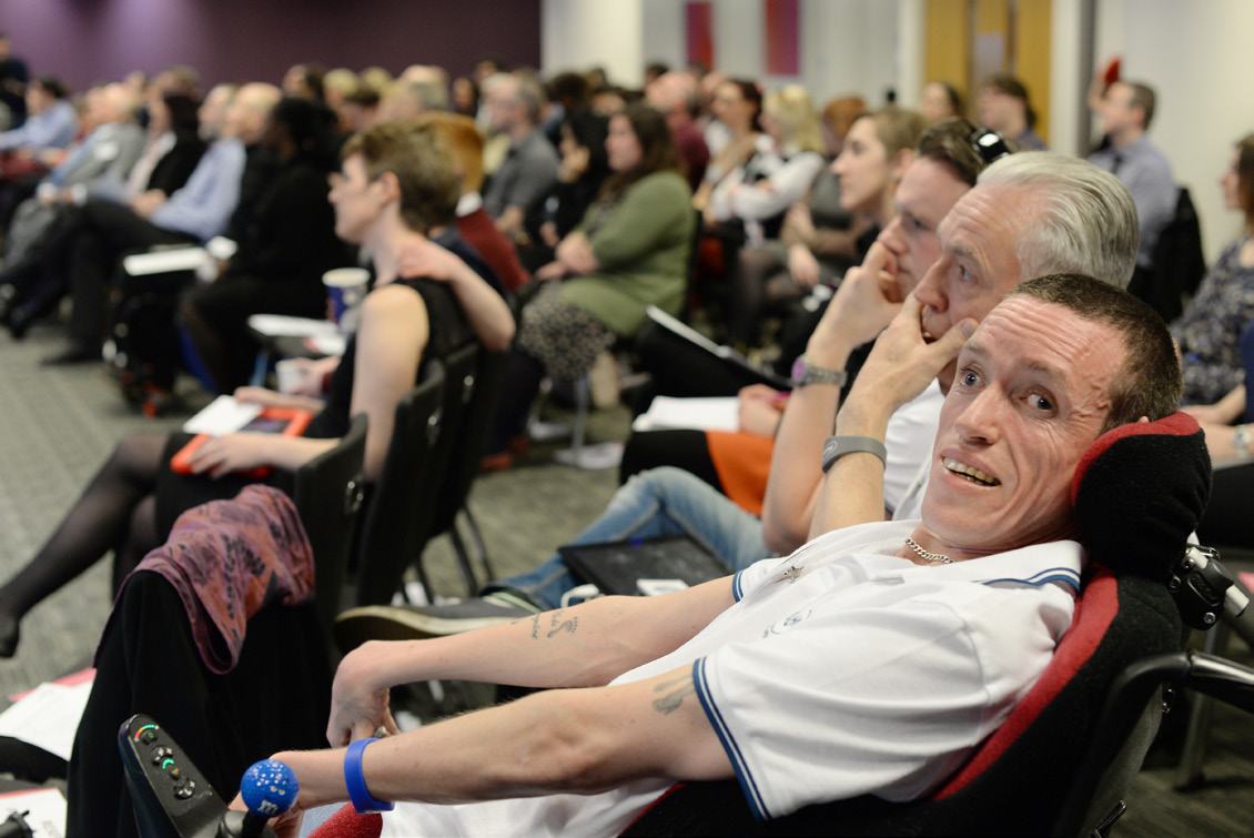 A man in a wheelchair in the foreground looks at the camera, while other attendees are seated in an audience at an event.