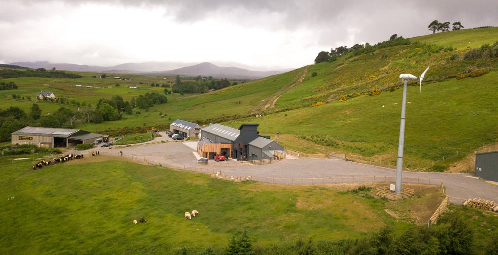 Aerial view of a modern building complex with a wind turbine on a green hillside under a cloudy sky.