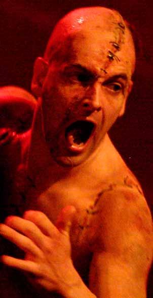 Close-up of a bald actor with stage makeup depicting stitches, shouting intensely under red light.