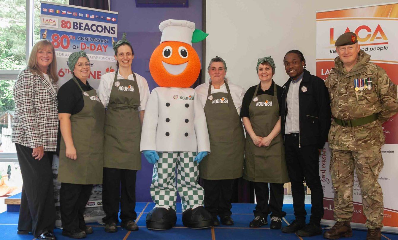 A group photo of eight people, including catering staff in aprons, an orange vegetable mascot, a man in military uniform, and others, posing at an event with "LACA" and "80th ANNIVERSARY of D-DAY" banners.
