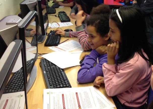 Two young girls smile while engaging with a computer in a classroom, illustrating a learning or technology education activity.