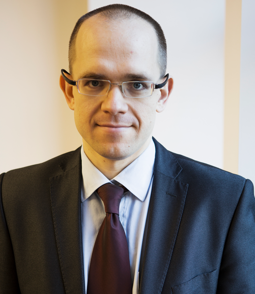 Headshot of a man with short brown hair, wearing glasses, a light blue shirt, dark suit, and maroon tie.