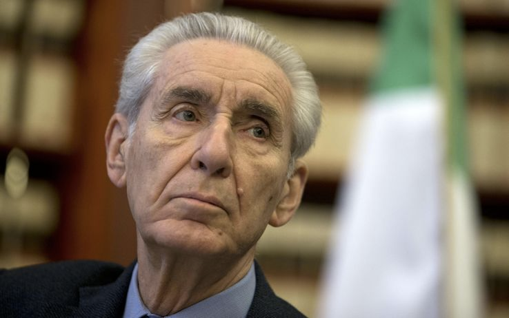 Close-up portrait of an older man with gray hair, looking to the right. An out-of-focus Italian flag is in the background.