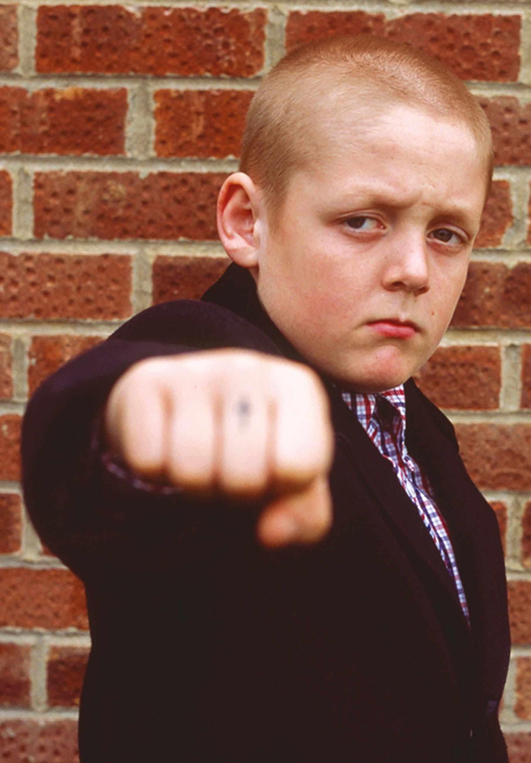 Young boy in a dark suit with a plaid shirt, making a fist towards the camera against a brick wall.