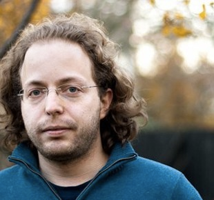 Headshot of a man with curly brown hair and glasses, wearing a blue jacket, looking forward.