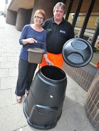 A man and a woman stand with a black compost bin and a smaller brown food waste bucket, promoting composting.