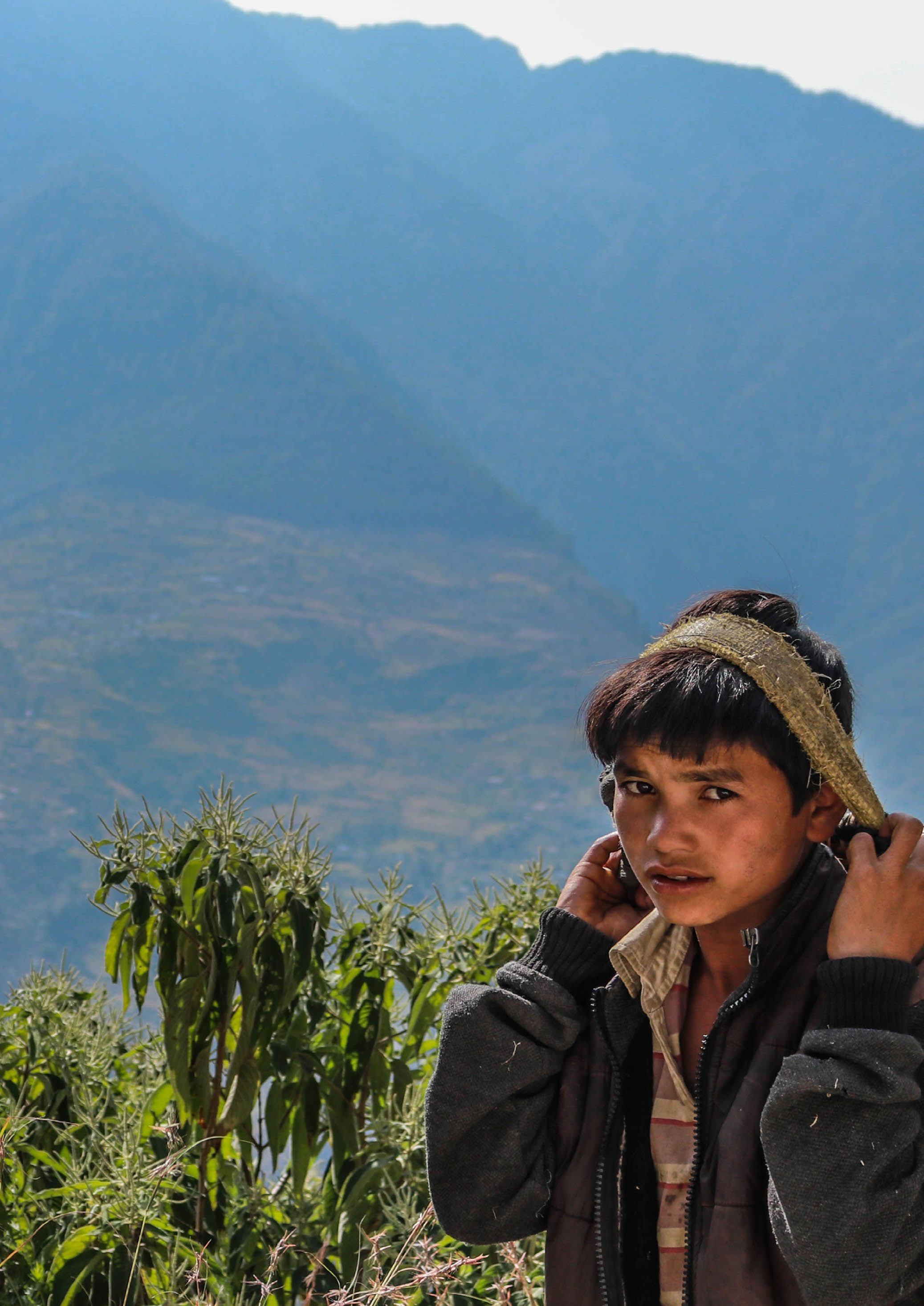A close-up portrait of a young boy with a cloth headband, wearing a dark jacket, against a background of green plants and mountains.