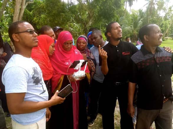 A group of people outdoors, some looking up, while a woman in the center holds a drone controller with an attached smartphone.