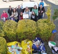 A group of adults and children stand behind bushes with large bags of collected trash in the foreground, indicating a community clean-up event.