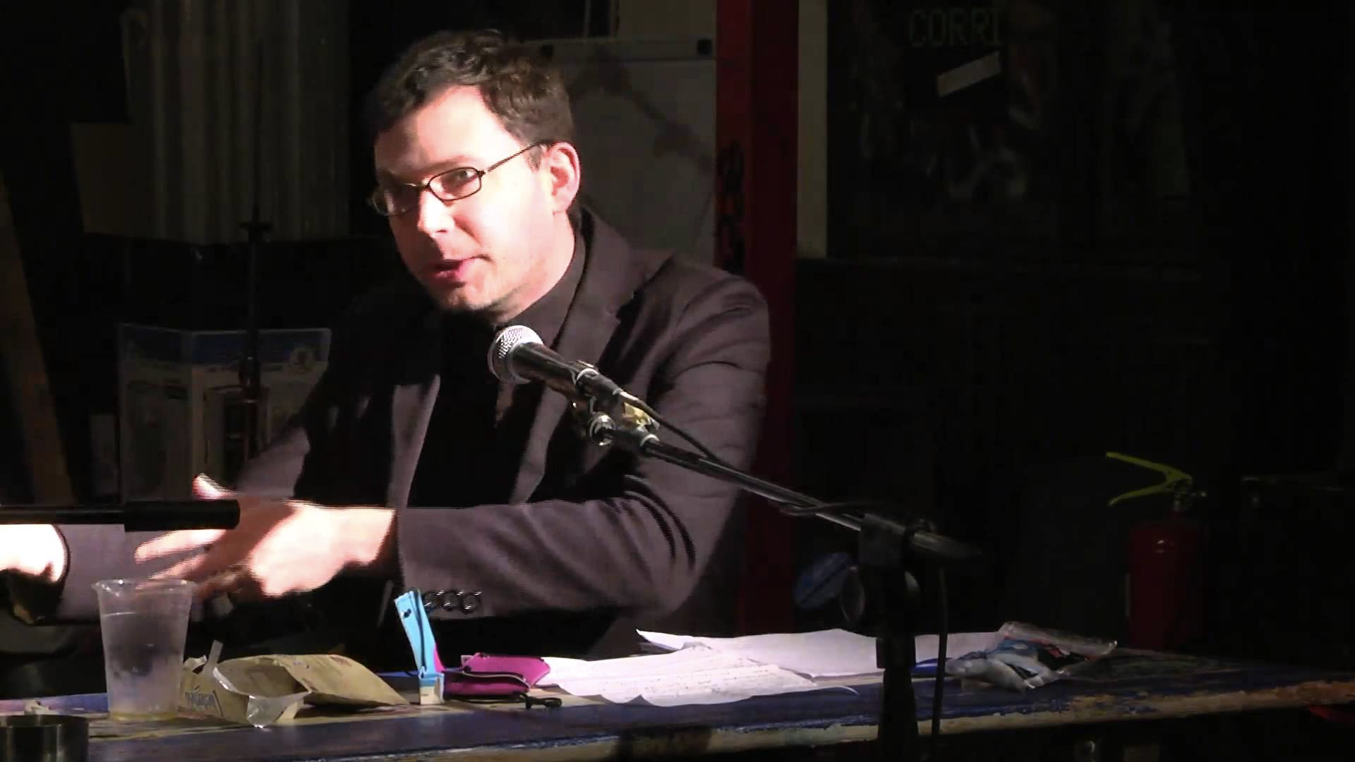 A man with glasses speaks into a microphone at a table, gesturing with his left hand during an event.