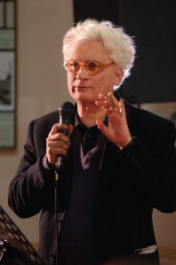A man with white curly hair and orange glasses speaks into a microphone, gesturing with his right hand.