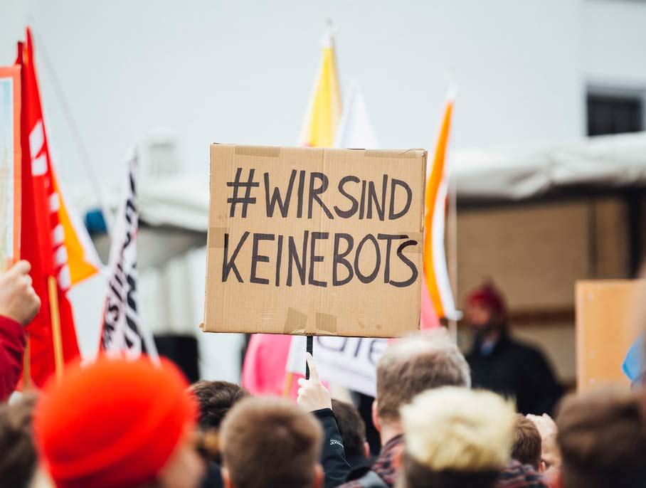A protestor holds a cardboard sign with '#WIRSIND KEINEBOTS' (We are not bots) written on it, surrounded by a crowd at an outdoor event.