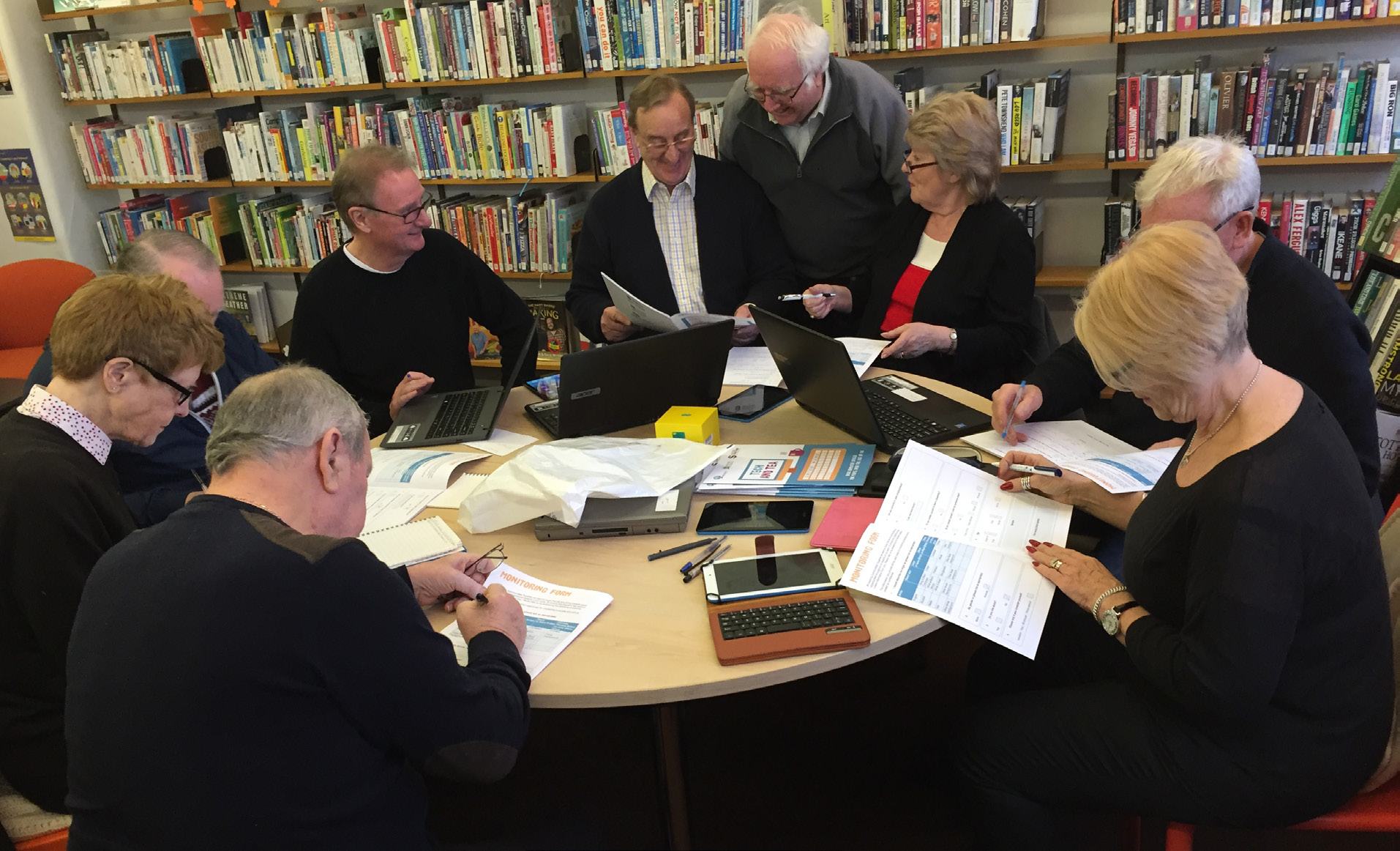 A group of older adults are gathered around a table in a library, engaged in a discussion or learning activity with laptops and papers.