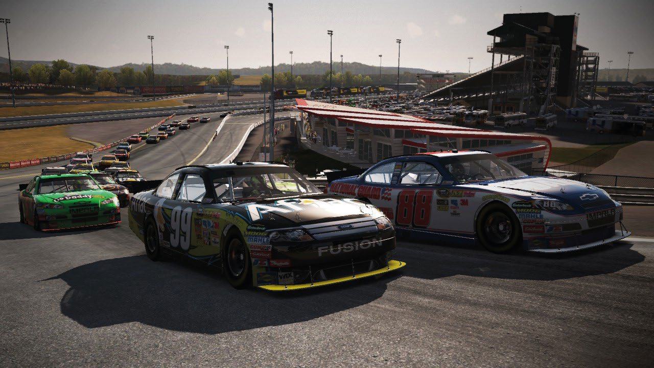 Several NASCAR race cars on a track, with grandstands and a large stadium structure visible in the background under a sunny sky.