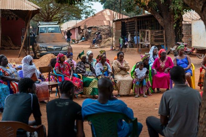Group of people, mainly women, sitting in a circle outdoors in a village, participating in a community meeting.