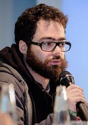 A man with a beard and glasses speaks into a microphone, focused on the audience.