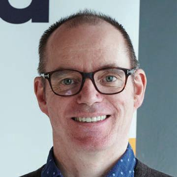 Headshot of a smiling man with short hair and glasses, wearing a blue patterned shirt.