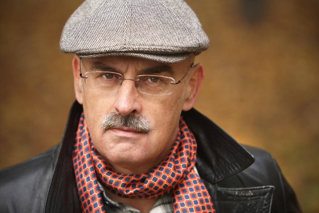 Portrait of a man with a mustache and glasses, wearing a gray flat cap, black jacket, and patterned scarf.