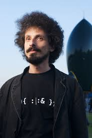 Portrait of a man with curly hair and beard, wearing a black t-shirt with symbols, standing outdoors near a rocket-shaped structure.