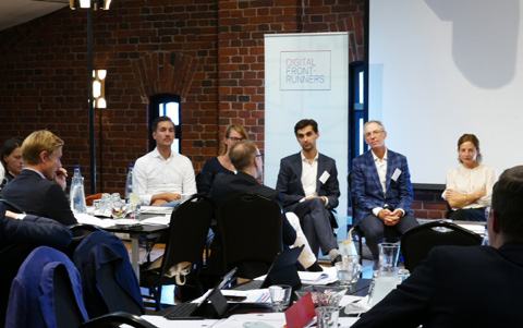 Individuals participate in a meeting or panel discussion with a "DIGITAL FRONT-RUNNERS" banner in the background.