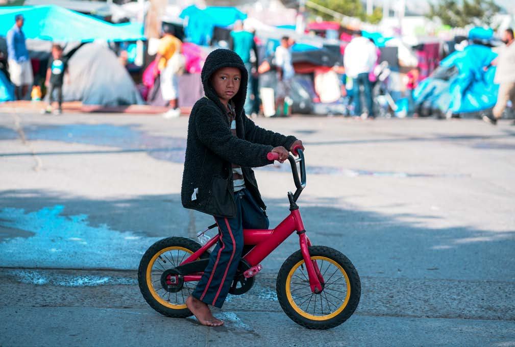 A barefoot child on a red bicycle in front of a refugee camp, looking at the camera, depicting childhood in a displacement setting.